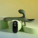 The Strap Tap & Go Girth Enhancer & Tapping Vibrator, a green silicone hands-free couples toy with an ergonomic shape, rests on light green cylinders beside its two-button black remote, against a soft yellow and green background.