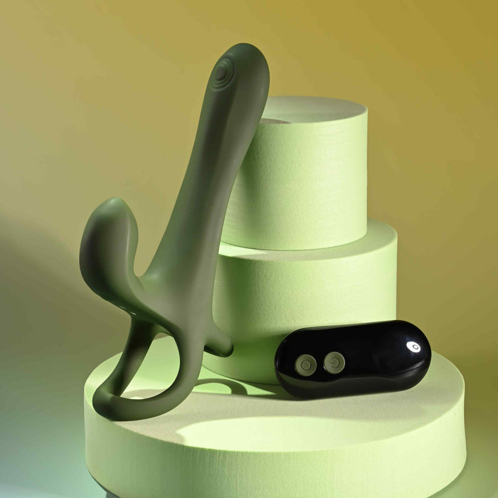 The Strap Tap & Go Girth Enhancer & Tapping Vibrator, a green silicone hands-free couples toy with dual stems, stands upright on pale green platforms beside its small black two-button remote. Soft yellow and green lighting complete the scene.