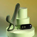 The Strap Tap & Go Girth Enhancer & Tapping Vibrator, a green silicone hands-free couples toy with dual stems, stands upright on pale green platforms beside its small black two-button remote. Soft yellow and green lighting complete the scene.