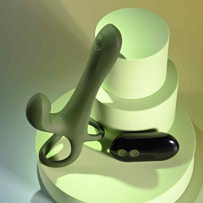 The Strap Tap & Go Girth Enhancer & Tapping Vibrator and its black remote control are shown on light green cylindrical platforms under soft, multicolored lighting.