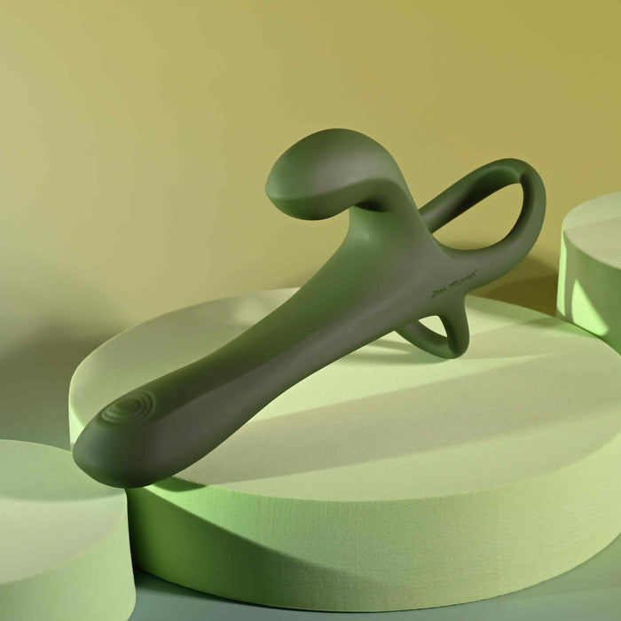 The Strap Tap & Go Girth Enhancer & Tapping Vibrator, a modern green silicone device with a curved design, is shown on circular pale green platforms against a soft yellow background.
