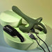 The Strap Tap & Go Girth Enhancer & Tapping Vibrator, a green silicone hands-free couples toy with USB charging cable, is shown on a round light green platform next to its black case against soft green and yellow gradients.