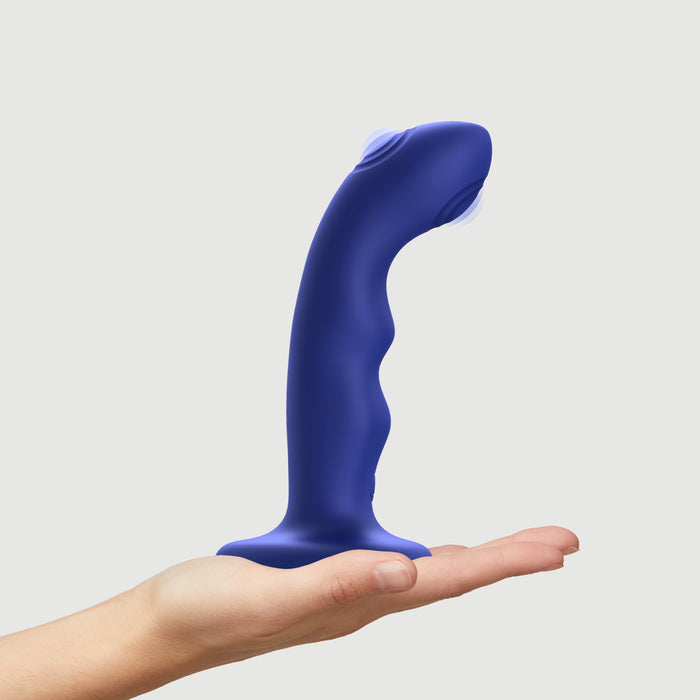 A hand holds the Strap on Me Curved G-Spot Tapping Blue Silicone Dildo, featuring a curved shape and flared base, isolated on a plain white background.