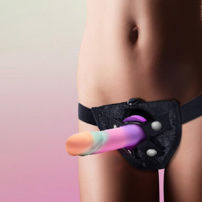 A person wears a black strap-on harness with the Sunrise Gaze Textured Silicone Suction Cup 7.5 Inch Dildo, featuring rainbow colors, standing against a pink and purple gradient background.