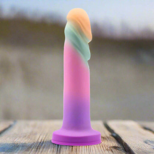 The Sunrise Gaze Textured Silicone Suction Cup 7.5 Inch Dildo—a pastel, column-shaped toy with a rounded swirled top—is upright on a wooden surface outdoors, designed for g-spot stimulation.