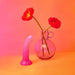 The Sunrise Smooth 7 Inch Pink Ombre Glitter Silicone Dildo with Suction Cup stands upright beside a vase of red poppies, set against a bright pink and orange background—ideal for prostate or g-spot stimulation.