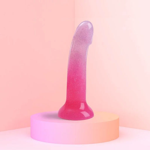 The Sunrise Smooth 7 Inch Pink Ombre Glitter Silicone Dildo with Suction Cup stands upright on a round platform, its sparkly silicone perfect for prostate or g-spot play, set against a pastel background.