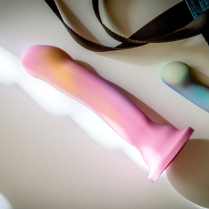 The Sun's Out 7.5 Inch Pink Ombre Silicone G Spot Dildo with a flared base rests on a white surface in sunlight, with a brown strap and another toy partially visible nearby.