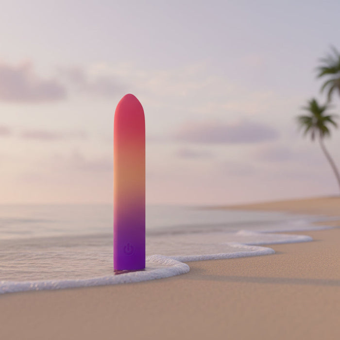 The Sunset Passion Soft Bendable Bullet Vibrator, with a pink and orange gradient, stands upright in wet shoreline sand as gentle waves roll in and a blurred palm tree appears beneath a pastel sky.
