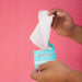 Two hands hold an open package of Sweetums Flavored Intimate Wipes, featuring a turquoise blue and white design against a pink background. Made from plant fibers, one hand smoothly pulls a single white wipe from the pH-balanced pack.