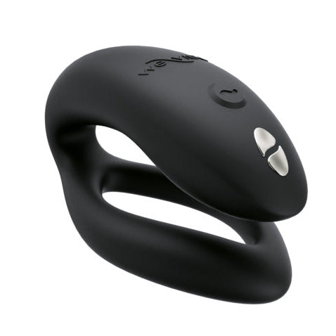 The Womanizer x We-Vibe Lust & Luxury Collection features a black, curved silicone couples vibrator with two silver buttons and an on/off symbol. Its ergonomic, modern design has two connected loops for intimate pleasure.