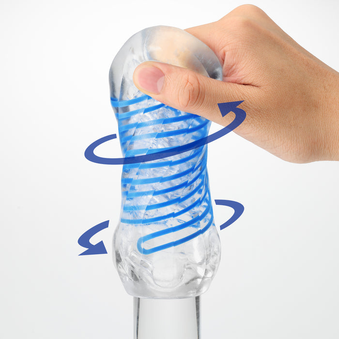 A hand squeezes a transparent Tenga Spinner DX Drops Textured Masturbator, showing its thicker, textured elastomer interior with blue spiral arrows that indicate the stroker’s spinning motion, all set against a plain white background.