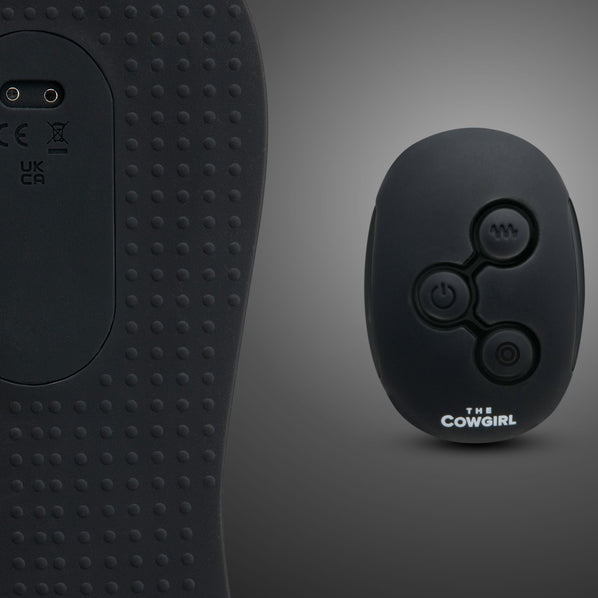 A close-up of a textured black surface with control buttons beside a small black oval remote labeled "The Cowgirl Rodeo Rumbler Grinding Sex Machine" with three buttons, designed for hands-free pleasure on a gray gradient background.