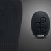 A close-up of a textured black surface with control buttons beside a small black oval remote labeled "The Cowgirl Rodeo Rumbler Grinding Sex Machine" with three buttons, designed for hands-free pleasure on a gray gradient background.