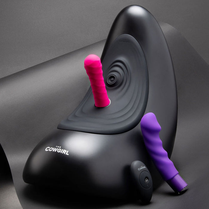 The Cowgirl Saddle Sex Machine with Vibration, Thrusting, and Rocking is shown in black with a pink ribbed attachment on top and a purple attachment beside it, designed for hands-free pleasure against a dark background.