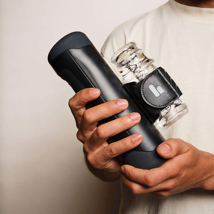 A person in a white shirt holds The Handy Adjustable Hand Job Penis Stroker, a sleek black interactive male masturbator with a small clear plastic container attached by a black strap.