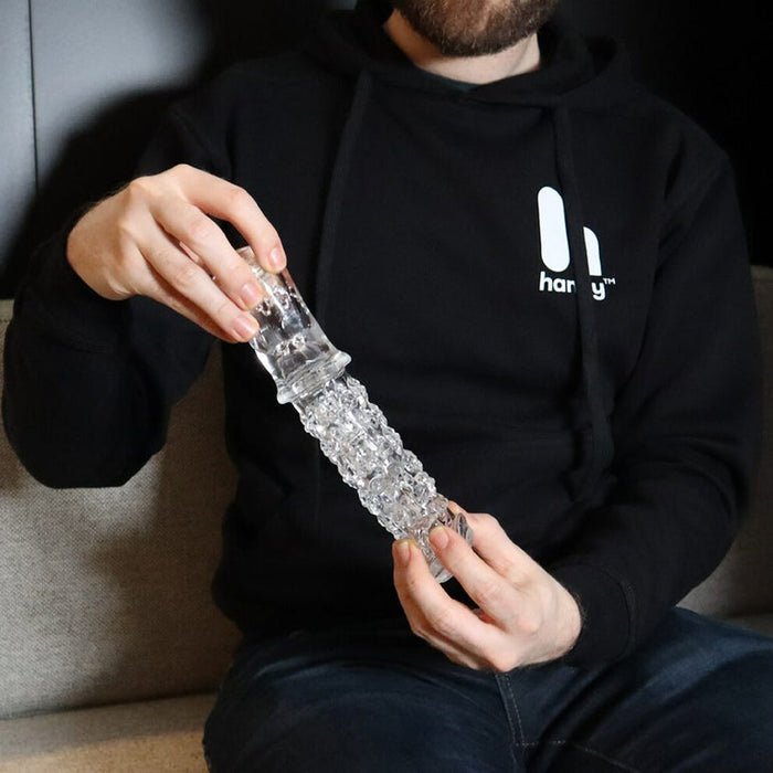 A person in a black hoodie sits on a couch, holding The Handy Dream Collection Chrysm Sleeve—a clear, textured stroker—with both hands. Their face is partially out of the frame.