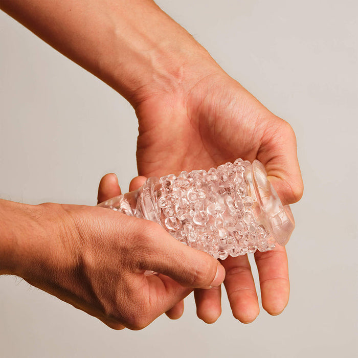 A person holds and squeezes The Handy Dream Collection Chrysm Sleeve—a transparent, textured stroker with bubble-like bumps—against a neutral background, designed to enhance anal penetration sensations.