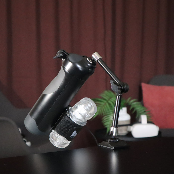 The Handy Handsfree Desk Mount holds a black water bottle and a small clear container with its adjustable articulating arm. In the softly blurred background, you can see a dark curtain, plant, and tech devices.