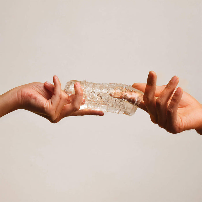 Two hands exchange The Handy Mermaid Collection Pearl Sleeve, a transparent, textured penis sleeve male masturbator with a bubble-like surface, set against a plain beige background.