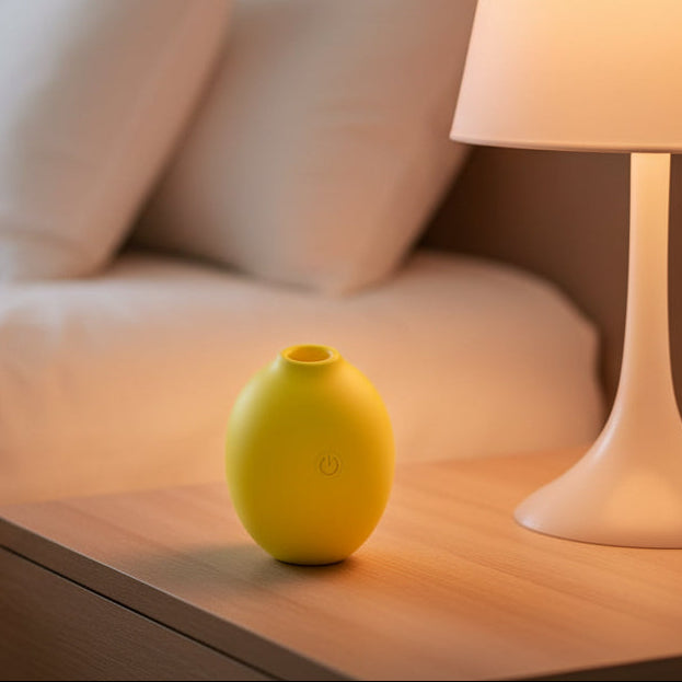 The Lemon Powerful Clitoral Air Pulsation Suction Vibrator, a small yellow oval device with a power button, rests discreetly on a wooden bedside table beside a white lamp, offering clitoral pleasure with a bed and pillow in the background.