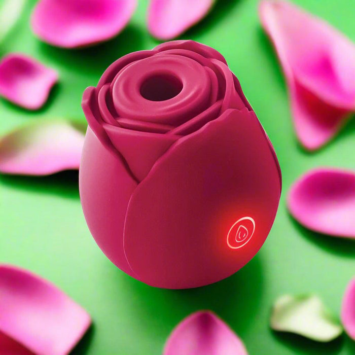 The Rose Powerful Clitoral Suction Air Pulsation Vibrator, a pink, rose-shaped silicone stimulator with a power button and red indicator light, is pictured among pink rose petals on a green background.