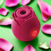 The Rose Powerful Clitoral Suction Air Pulsation Vibrator, a pink, rose-shaped silicone stimulator with a power button and red indicator light, is pictured among pink rose petals on a green background.