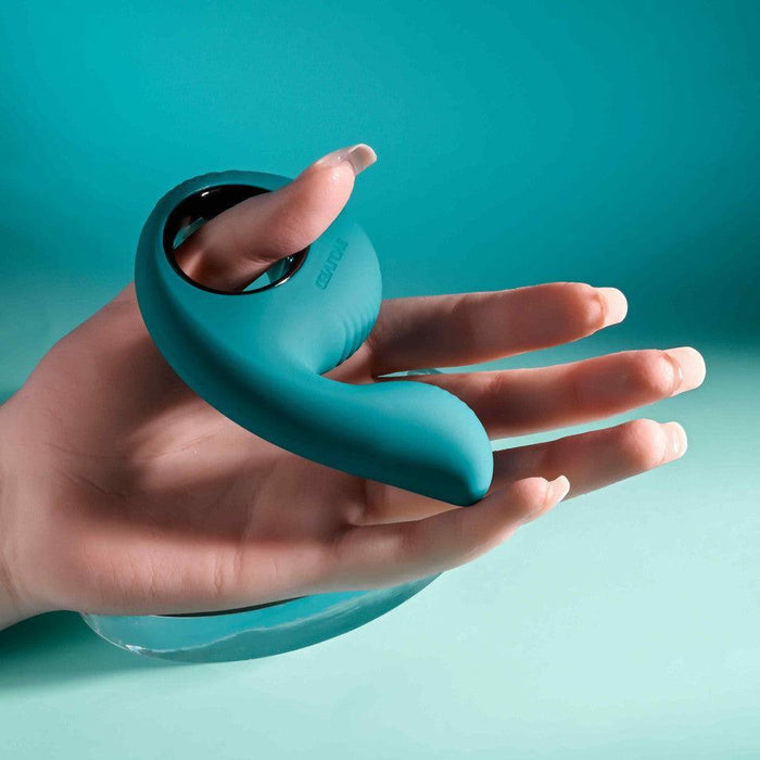 Teal silicone thumb-shaped vibrator with a finger loop held in a palm against a matching teal background.