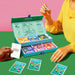 A person in a yellow top holds a die next to the open Together Couples Intimacy Card Game box with illustrated cards and dice. Another hand has a white card. Cards labeled "Whisper," "Guess," and "Touch" inspire communication for couples.