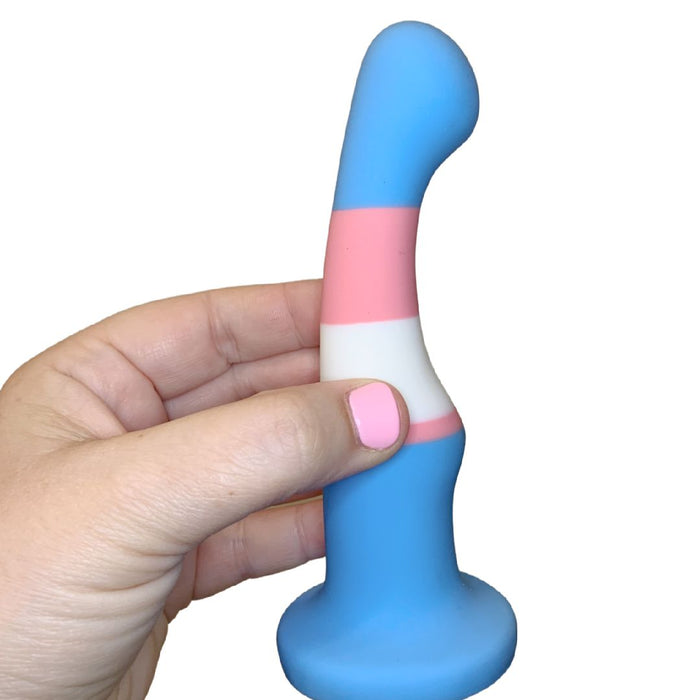 A hand with pink nail polish holds the Trans Pride Slim Curved 6 Inch Silicone Suction Cup Dildo, featuring blue, pink, and white stripes inspired by the transgender pride flag.