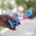A person with purple-painted nails holds the Trans Pride Slim Curved 6 Inch Silicone Suction Cup Dildo, featuring blue, pink, and white trans pride colors, a flared base, and curved tip. The background is blurred with "blush" in the lower right corner.