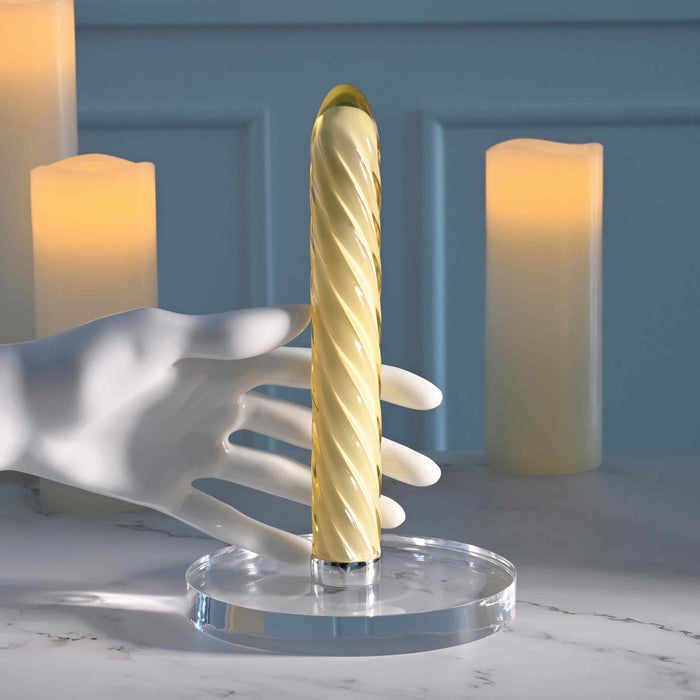 The Twist of Fate Slim Waterproof Rechargeable Glass Vibrator, a sculpted pale yellow piece with a spiral design, stands on a clear round base beside a white mannequin hand. Three flameless candles glow softly on the marble surface.