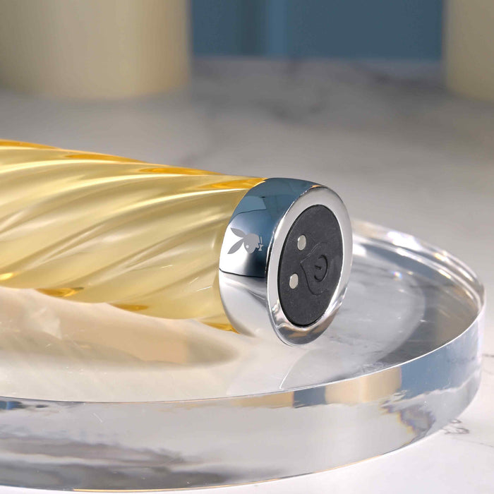 A close-up of the Twist of Fate Slim Waterproof Rechargeable Glass Vibrator, featuring a gold spiral design with a silver cap bearing the Playboy bunny logo, displayed on a clear round tray—an elegant choice for temperature play.