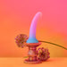 The Utopia Sleek 7 Inch Pink and Blue Ombre Silicone Dildo with Suction Cup stands upright on an ornate metallic pedestal, flanked by two spherical purple flowers, against a vivid pink and orange gradient background.