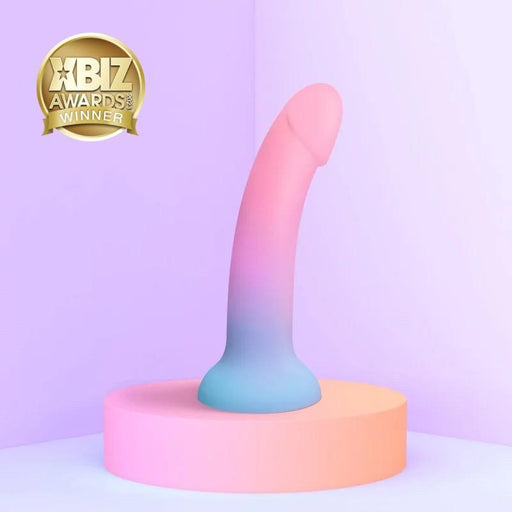 The Utopia Sleek 7 Inch Pink and Blue Ombre Silicone Dildo with Suction Cup stands on a round platform in a softly lit, minimal room. An "XBIZ Awards Winner" gold badge appears at the top left—ideal for prostate or G-spot play.