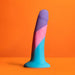 The Vision of Love Tapered 5.5 Inch Silicone Suction Cup Dildo, crafted from platinum silicone in blue, pink, and purple hues, stands upright on an orange background—an ideal choice for beginners.