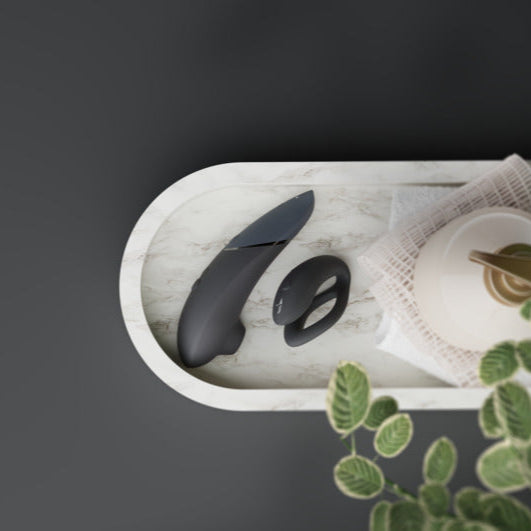 The Womanizer x We-Vibe Lust & Luxury Collection couples vibrator and matching remote rest on a white marble tray beside a teapot and napkin, with green leaves in the foreground.