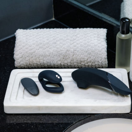 A marble tray on the bathroom counter holds three black massagers from the Womanizer x We-Vibe Lust & Luxury Collection. Behind the tray are a rolled white towel and a bottle of clear liquid with a black cap.