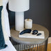 A modern bedside table holds a white lamp, a textured ceramic jar with gold knob, and three black massagers from the Womanizer x We-Vibe Lust & Luxury Collection on a marble tray; part of a bed with white linens and a green pillow is visible.