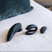 The Womanizer x We-Vibe Lust & Luxury Collection—featuring three sleek black personal massagers and a small remote—is displayed on a neatly made bed with white striped sheets and a green pillow in the background.