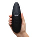 A hand holds the sleek, black Womanizer x We-Vibe Lust & Luxury Collection device with three round buttons and two small indicator lights, designed for couples’ pleasure with innovative Pleasure Air technology.