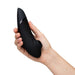 A hand holds the sleek, black Womanizer x We-Vibe Lust & Luxury Collection with a curved design and round opening, highlighting its Pleasure Air technology against a plain white background.