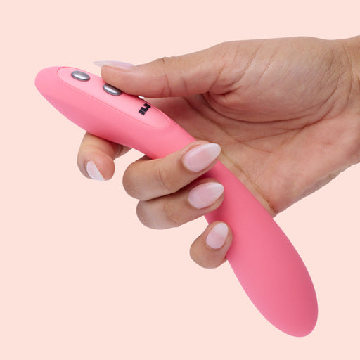 A hand holds the Ily Flexible Warming G-Spot Vibrator, a pink curved wand with buttons, made from body-safe silicone, against a light pink background.
