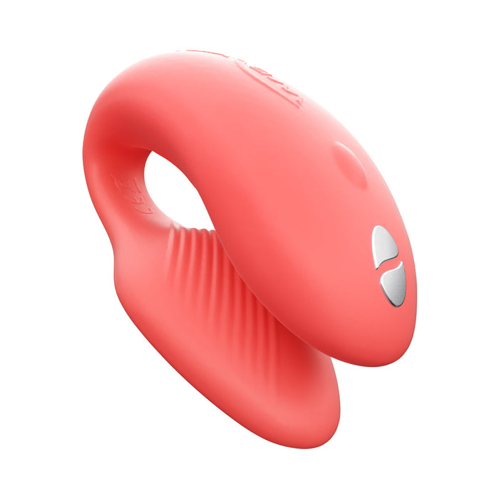 The We-Vibe Chorus Remote & App Controlled Couples' Vibrator in Coral is a pink, curved silicone couples toy with an adjustable fit, smooth finish, ribbed inner surface, and two silver buttons, shown on a white background.