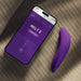 A smartphone with the We-Vibe Melt 2 Rechargeable Pleasure Air Clitoral Stimulator app is beside the curved, waterproof purple device on striped fabric, both shown in gentle natural light.