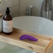 A wooden bath tray holds a We-Vibe Melt 2 Rechargeable Pleasure Air Clitoral Stimulator in purple and a brown liquid soap bottle, with a white bathtub and faucet visible in the background.