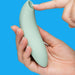 A hand holds the mint-green We-Vibe Melt 2 Rechargeable Pleasure Air Clitoral Stimulator with its curved, waterproof silicone body, while another hand presses the tip's opening, set against a bright blue background.
