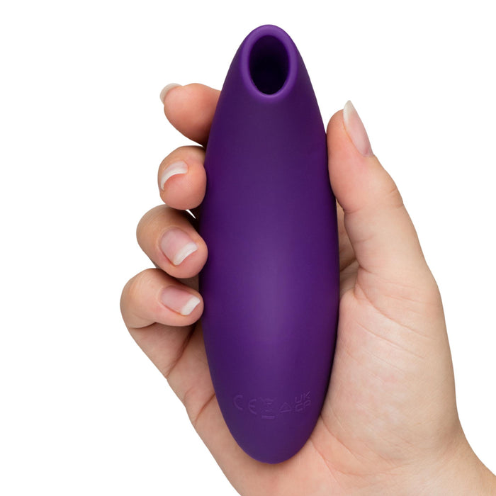 A hand holds the We-Vibe Melt 2 Rechargeable Pleasure Air Clitoral Stimulator, a purple, oval-shaped silicone device with a rounded tip and small opening, designed for air clitoral stimulation; regulatory symbols are embossed near its base.