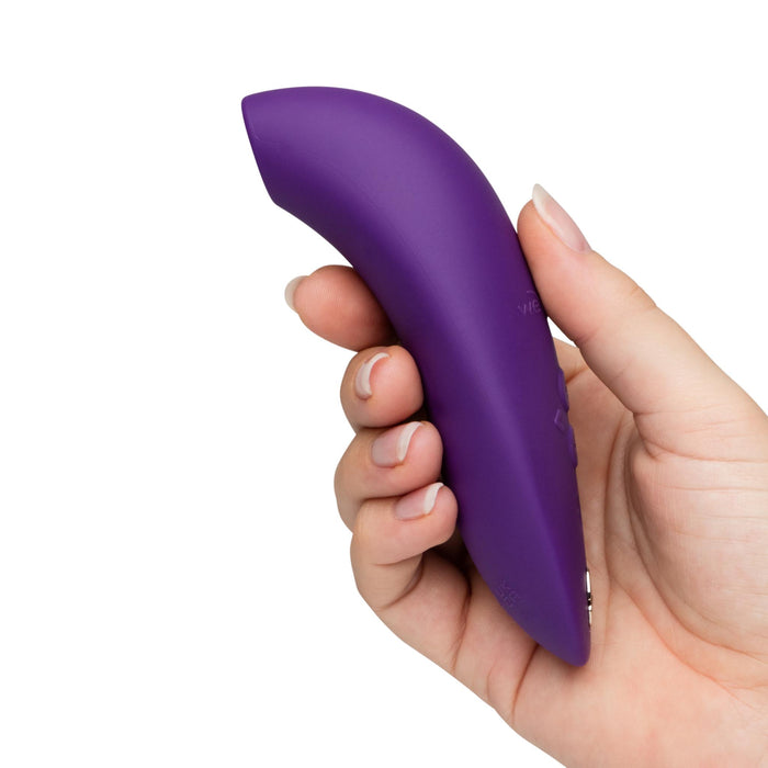 A hand holds the We-Vibe Melt 2 Rechargeable Pleasure Air Clitoral Stimulator, a purple, curved silicone massager with a waterproof body, against a plain white background.