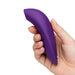 A hand holds the We-Vibe Melt 2 Rechargeable Pleasure Air Clitoral Stimulator, a purple, curved silicone massager with a waterproof body, against a plain white background.